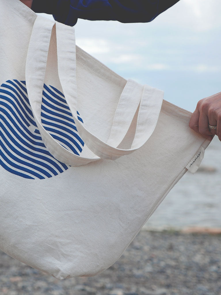 Sac tote bag (sac de marché) tenu par une main sur fond de plage du bas du fleuve. Sac en coton épais couleur naturelle avec logo sein-vague et étiquette Marées.