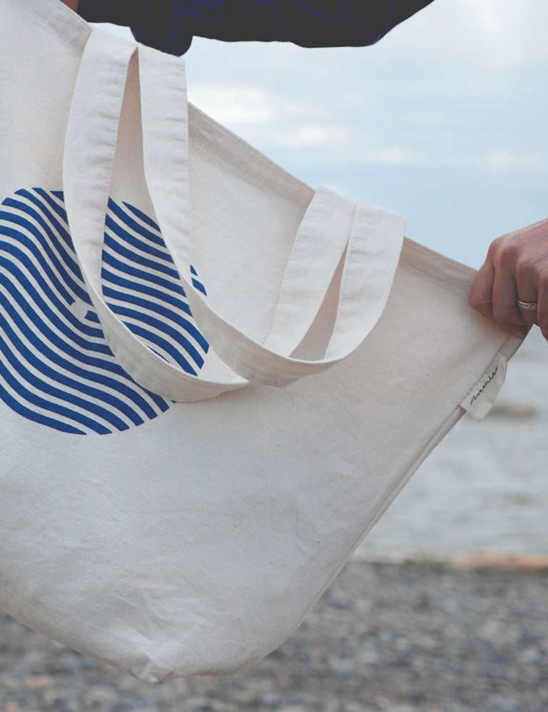 Sac tote bag (sac de marché) tenu par une main sur fond de plage du bas du fleuve. Sac en coton épais couleur naturelle avec logo sein-vague et étiquette Marées.