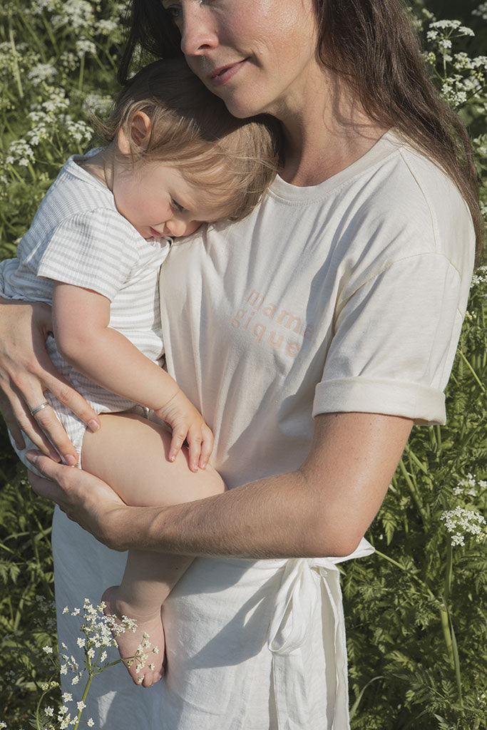 Maman dans un champ vert et fleuri qui tient sa fille de 21 mois dans ses bras et qui porte son son tee-shirt d’allaitement chandail Marées couleur Beige doux, avec des fermeture éclair latérales. Écriture sur le chandail d’allaitement rose pâle.  