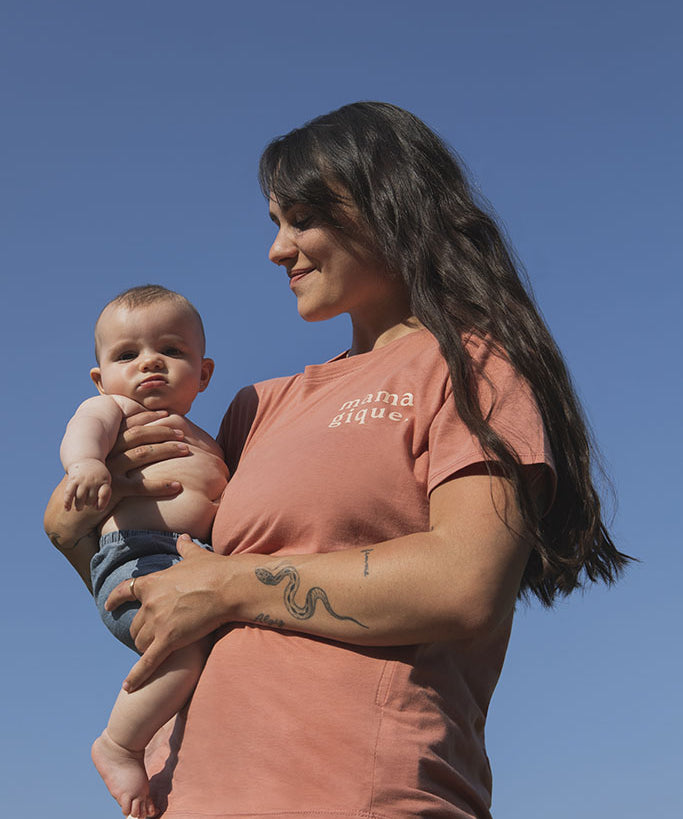 Maman qui tient son bébé de 5 mois dans ses bras et qui porte un tee-shirt, chandail d’allaitement Mamagique de Marées couleur Terra. Sur fond de ciel bleu en contre-plongée. Fermeture éclair latérales, coton biologique. Écriture sur le chandail d’allaitement rose pâle. 