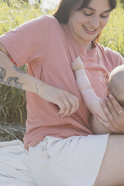 Maman avec son bébé de 5 mois qui s’apprête à allaiter sur une couverture crème lignée et qui porte un tee-shirt, chandail d’allaitement Mamagique de Marées couleur Terra. Fermeture éclair latérales, coton biologique. Écriture sur le chandail d’allaitement rose pâle. 