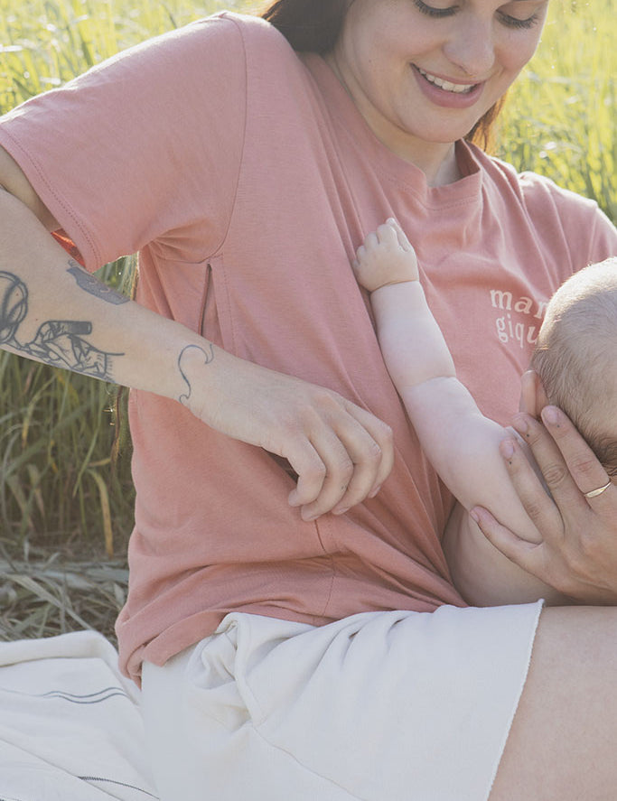 Maman avec son bébé de 5 mois qui s’apprête à allaiter sur une couverture crème lignée et qui porte un tee-shirt, chandail d’allaitement Mamagique de Marées couleur Terra. Fermeture éclair latérales, coton biologique. Écriture sur le chandail d’allaitement rose pâle. 