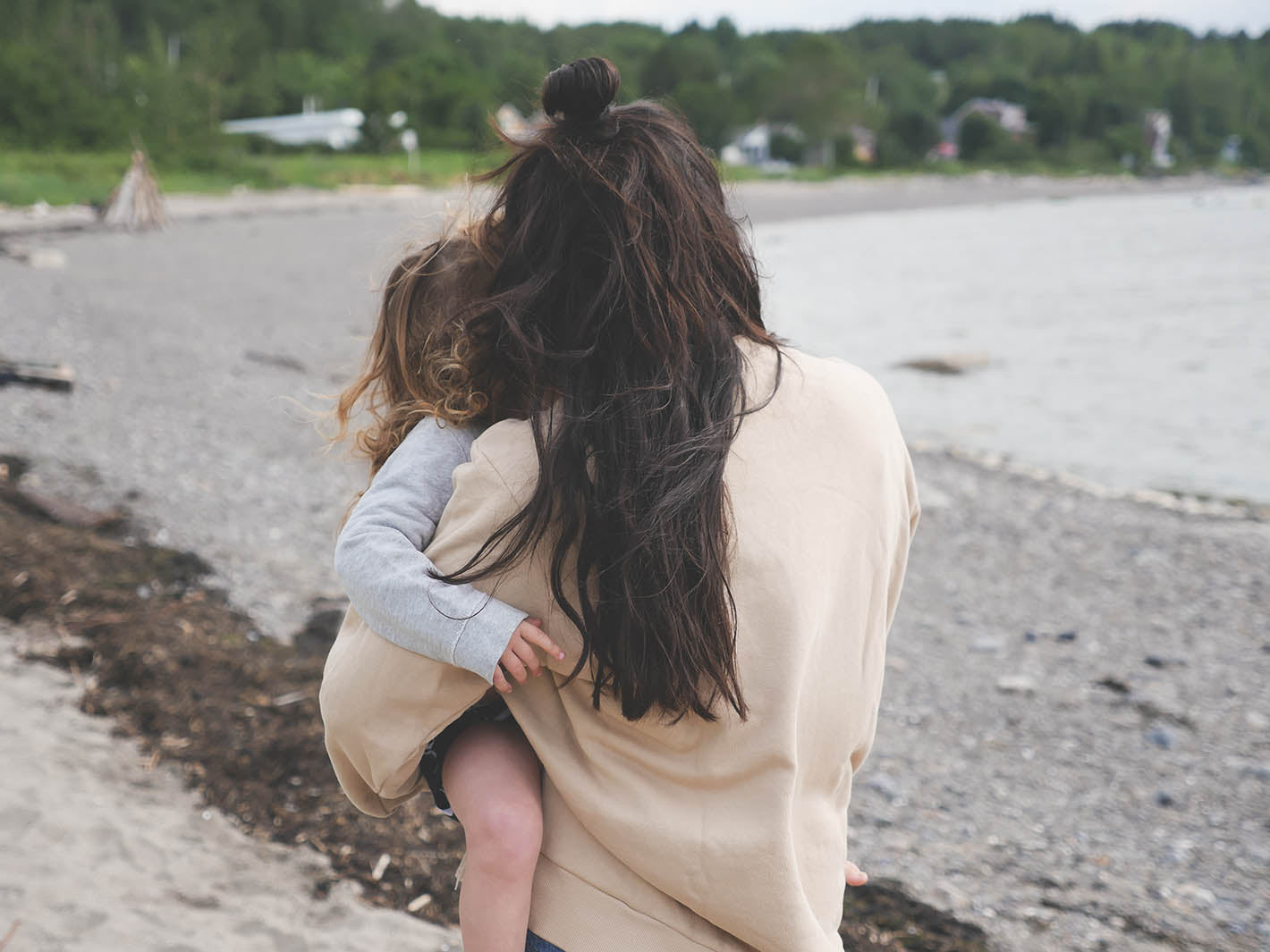 Maman de dos sur une plage au Québec, tient petite fille dans ses bras. Sweatshirt d'allaitement couleur sable doux. 