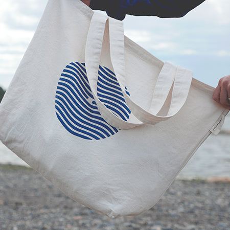 Sac type tote bag (sac de marché) tenu par deux mains sur fond de plage du bas du fleuve. Sac en coton épais couleur naturelle avec logo sein-vague et étiquette Marées. 