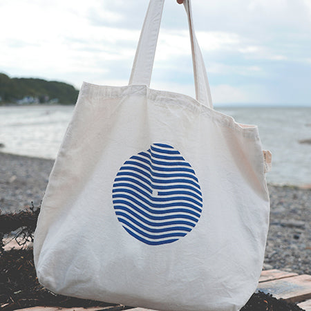 Sac type tote bag (sac de marché) tenu par une main sur fond de plage du bas du fleuve. Sac en coton épais couleur naturelle avec logo sein-vague et étiquette Marées.
