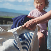 Sac type tote bag (sac de marché) en canevas de coton nature avec le logo sein vague. En arrière plan, on voir une fillette de 2 ans qui regarde ce que maman sort de son sac. Elle sort un Sweatshirt marine Marées. 