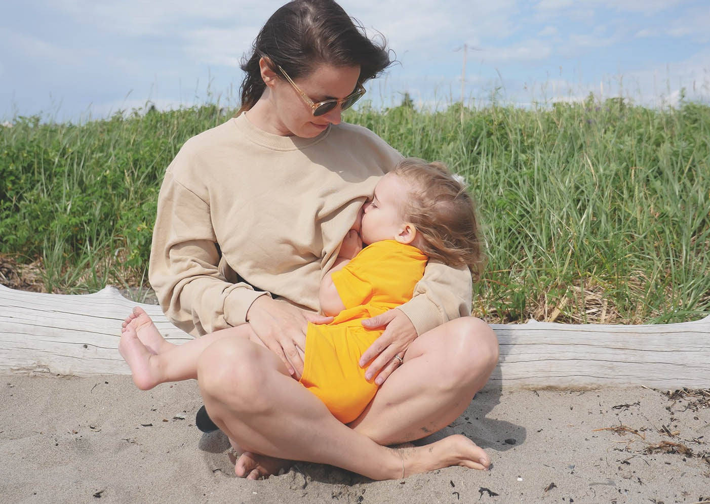 Maman qui allaite sa petite fille. Maman porte le sweatshirt d'allaitement Marées couleur sable. Elle est assis dans le sable sur une plage du bas du fleuve. Québec. 