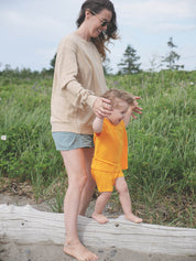 Maman qui port le sweatshirt d'allaitement Marées couleur sable. Elle fait marcher sa petite fille sur un tronc d'arbre sur une plage du Québec.