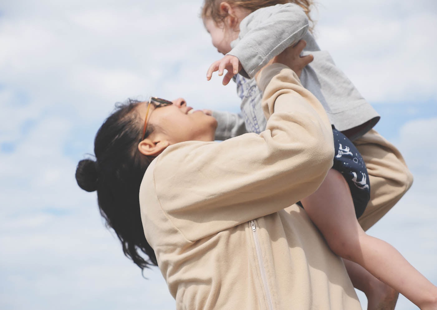 Maman qui porte le Sweatshirt d'allaitement Marées, couleur beige. La maman est de profil et elle tient sa petite fille dans ses bras. Derrière, on voit le ciel. Au Québec.