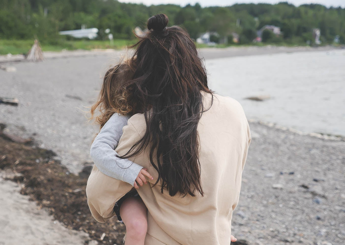 Maman qui porte le Sweatshirt d'allaitement Marées, couleur beige. La maman est de dos et elle tient sa petite fille dans ses bras. On voit une plage du bas du fleuve derrière. Au Québec.