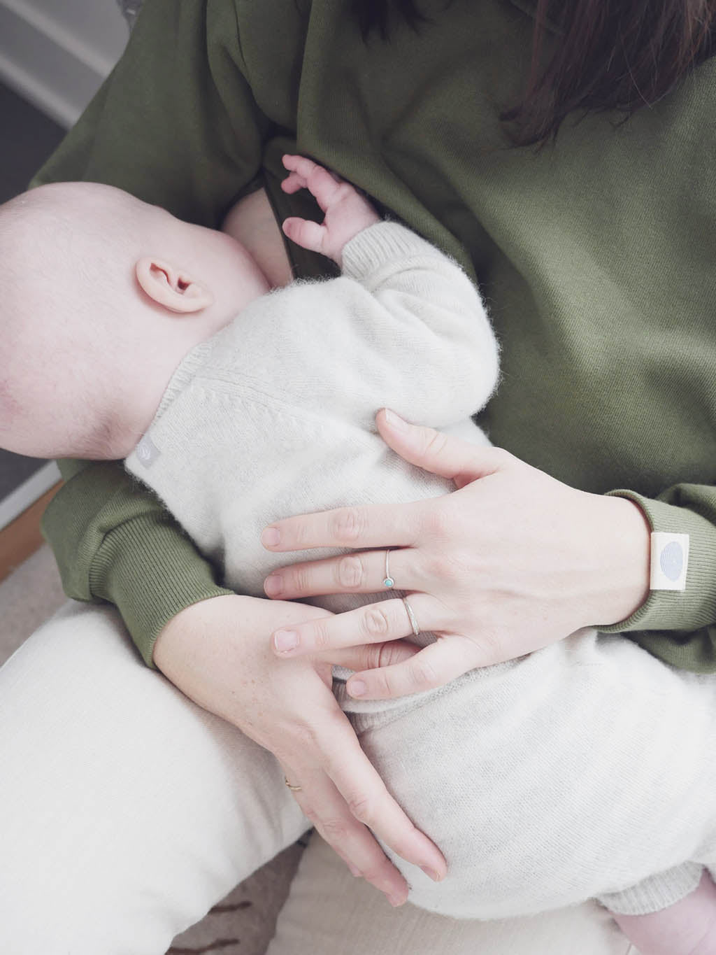 Gros plan sur Maman assise contre une fenêtre, qui tient bébé dans ses bras, avec son sweathshirt d’allaitement Marées, couleur seaweed. Bébé boit au sein dans ses bras.