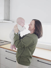 Maman dans la cuisine qui tient bébé dans ses bras devant elle et qui porte le sweatshirt d’allaitement Marées couleur seaweed.   
