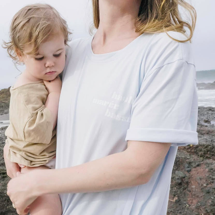 Gros plan femme tenant son bébé et portant le tee shirt d'allaitement maree haute maree basse bleu. Sur fond de plage. Rocher et vagues.
