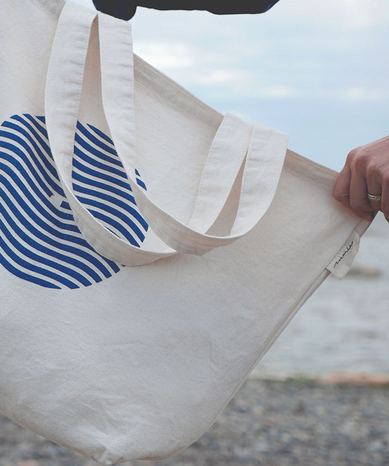 Sac tote bag (sac de marché) tenu par une main sur fond de plage du bas du fleuve. Sac en coton épais couleur naturelle avec logo sein-vague et étiquette Marées.
