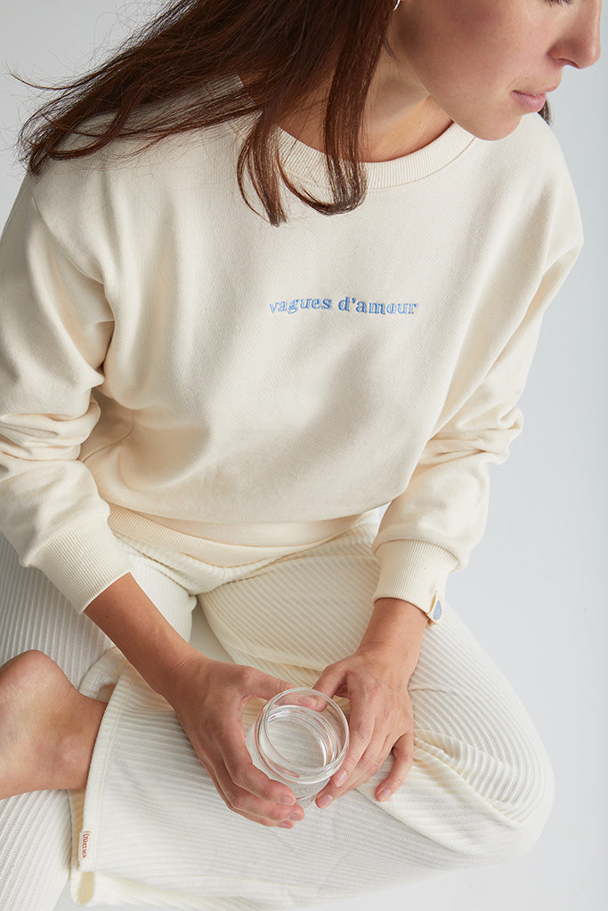 Maman assise boit un verre d'eau et porte le crewneck d'allaitement Vagues d'amour couleur nacre avec broderie bleue