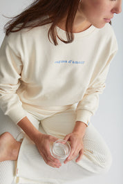 Maman assise boit un verre d'eau et porte le crewneck d'allaitement Vagues d'amour couleur nacre avec broderie bleue