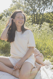 Maman et sa fille cachée derrière elle de 21 mois dans un champ avec des fleurs assises sur une couverte. Maman porte le tee-shirt d’allaitement chandail Marées couleur Beige doux avec des fermeture éclair latérales. Écriture sur le chandail d’allaitement rose pâle.