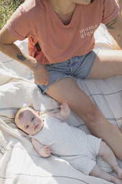 Maman avec son bébé de 5 mois sur une couverte crème lignée et qui porte un tee-shirt, chandail d’allaitement Mamagique de Marées couleur Terra. Fermeture éclair latérales, coton biologique. Écriture sur le chandail d’allaitement rose pâle. Maman s’apprête à allaiter. 