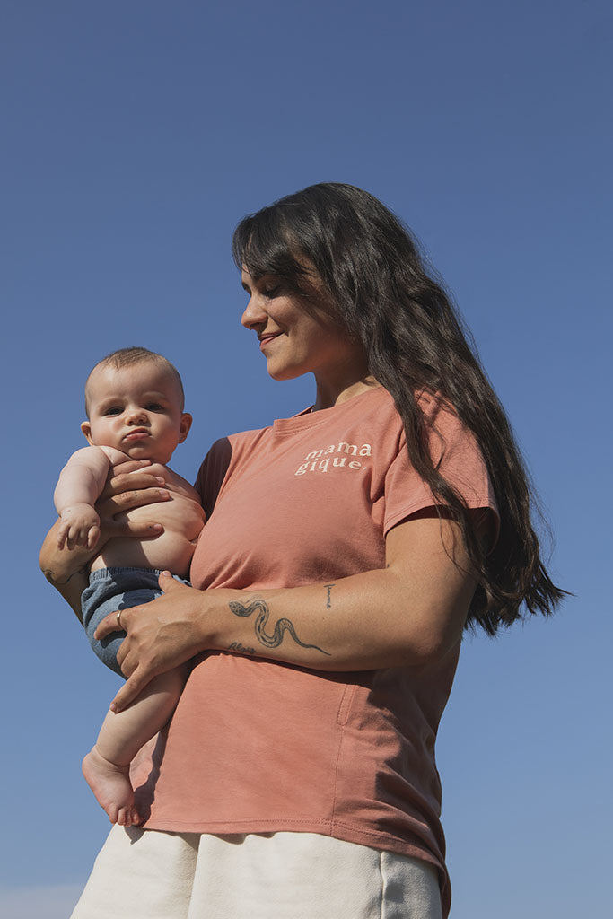 Maman qui tient son bébé de 5 mois dans ses bras et qui porte un tee-shirt, chandail d’allaitement Mamagique de Marées couleur Terra. Sur fond de ciel bleu en contre-plongée. Fermeture éclair latérales, coton biologique. Écriture sur le chandail d’allaitement rose pâle. 