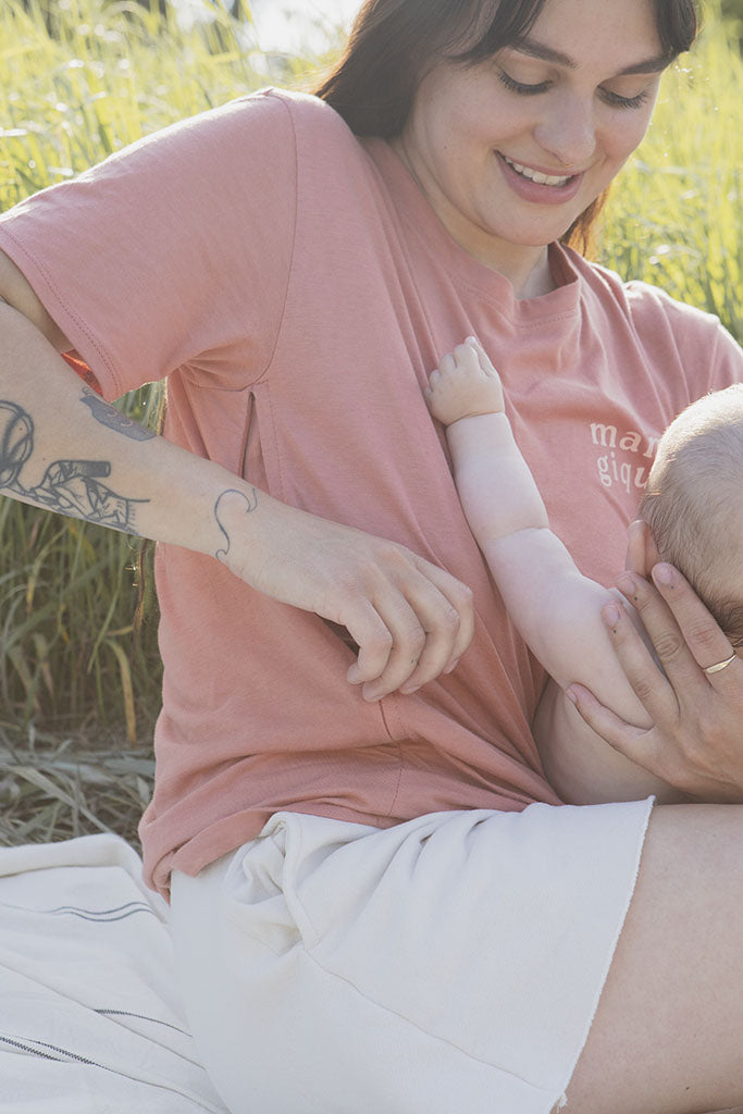 Maman avec son bébé de 5 mois qui s’apprête à allaiter sur une couverture crème lignée et qui porte un tee-shirt, chandail d’allaitement Mamagique de Marées couleur Terra. Fermeture éclair latérales, coton biologique. Écriture sur le chandail d’allaitement rose pâle. 