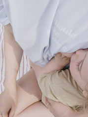 Plan serré sur femme assise sur la plage avec bébé au sein. Maman porte le tee shirt d'allaitement marée haute marée basse, bleu, sur un paréo blanc à rayures rouges.