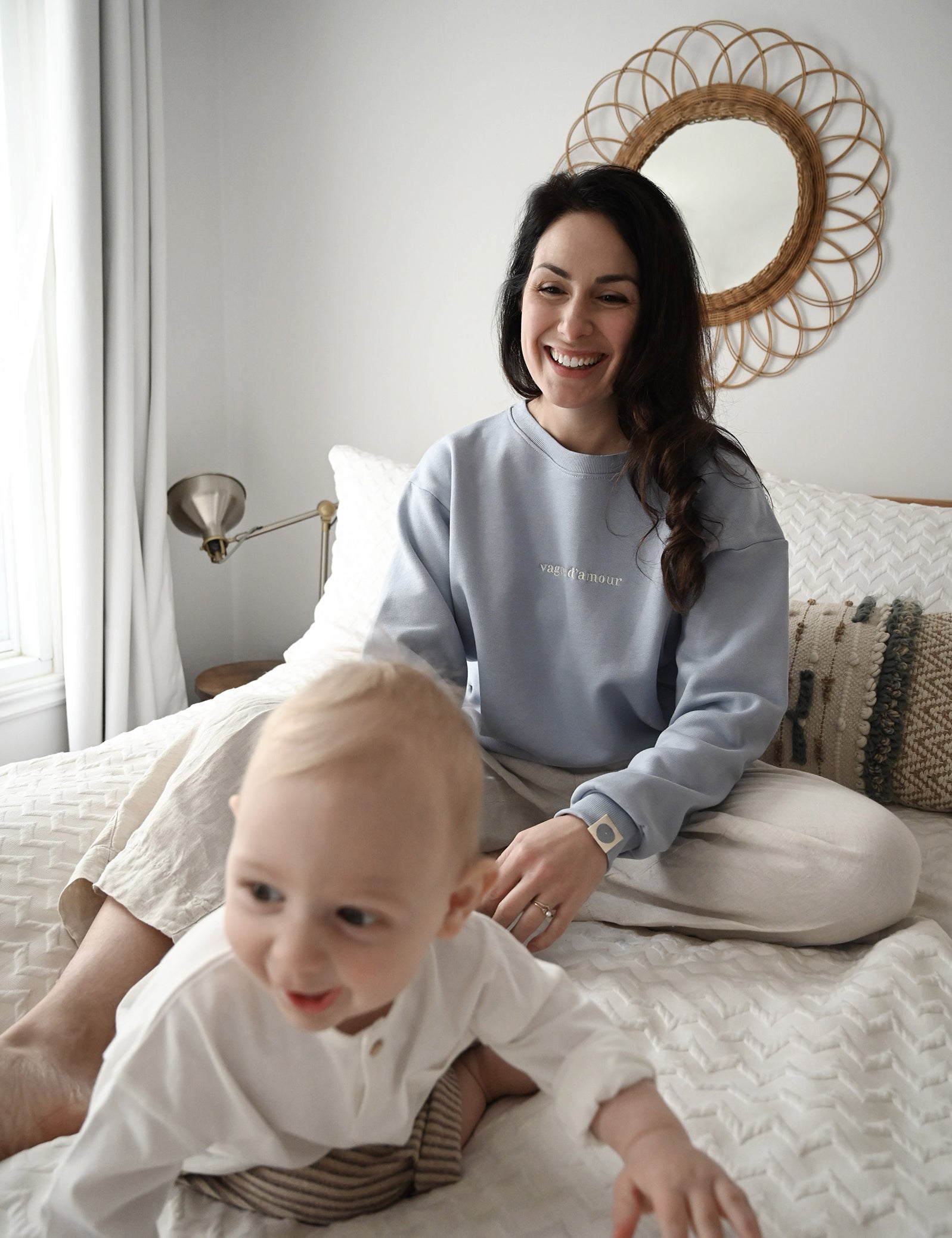 Maman sur son lit avec bébé qui sourrit. Mama porte le crewneck d'allaitement Marées bleu ciel. 