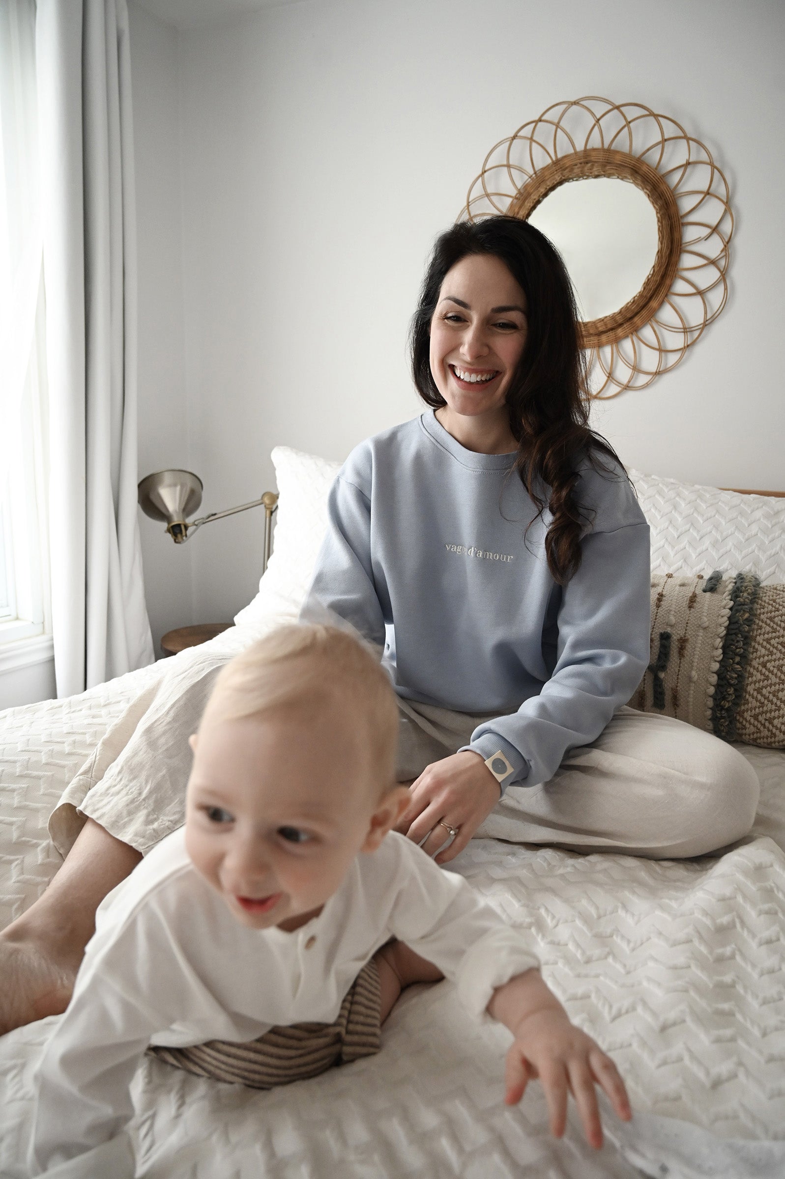 Maman sur son lit avec bébé qui sourrit. Mama porte le crewneck d'allaitement Marées bleu ciel. 