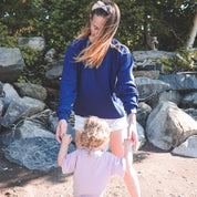 Maman qui porte le sweatshirt d'allaitement Marées marine et qui tient sa petite fille par les mains pour la faire tourner. Sur une plage du Québec.